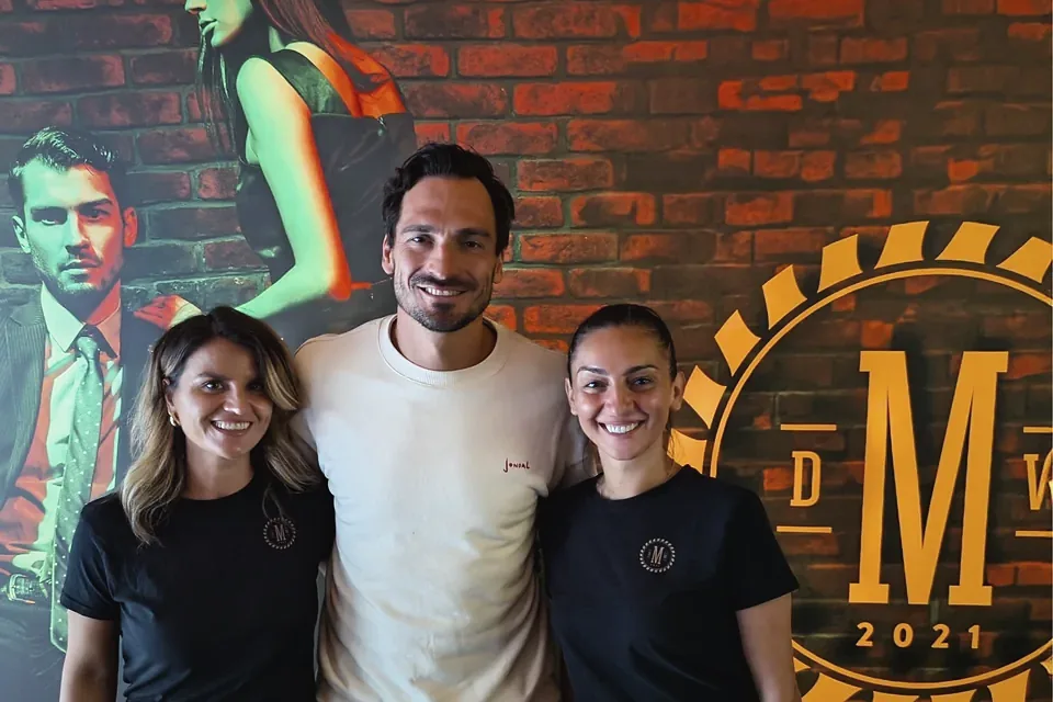 Mats Hummels mit zwei Kosmetikerinnen der Männer Werkstatt vor der Backsteinwand